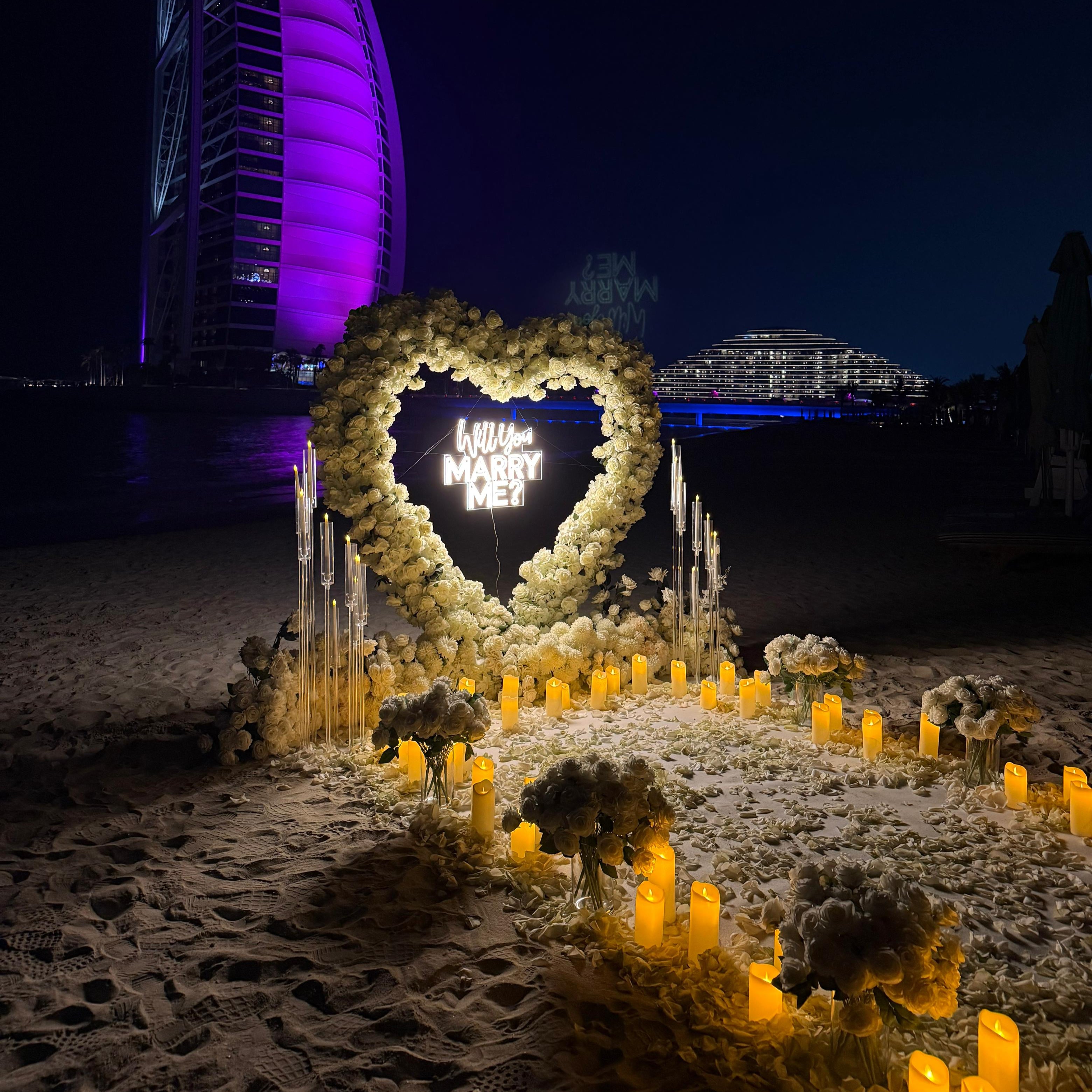 Heart-shaped installation with candles on a beach at night, with a purple-lit building in the background.